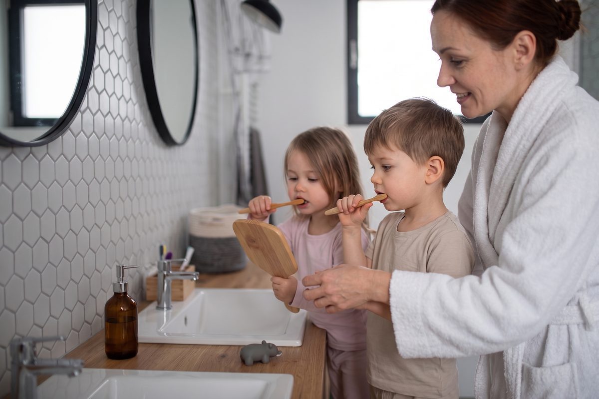 Madre con bata sostiene un espejo mientras dos niños se cepillan los dientes en el baño