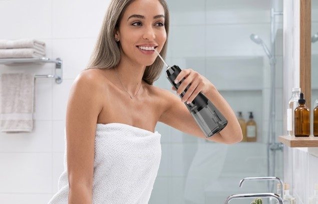 Mujer envuelta en toalla usa un irrigador oral negro frente al espejo del baño
